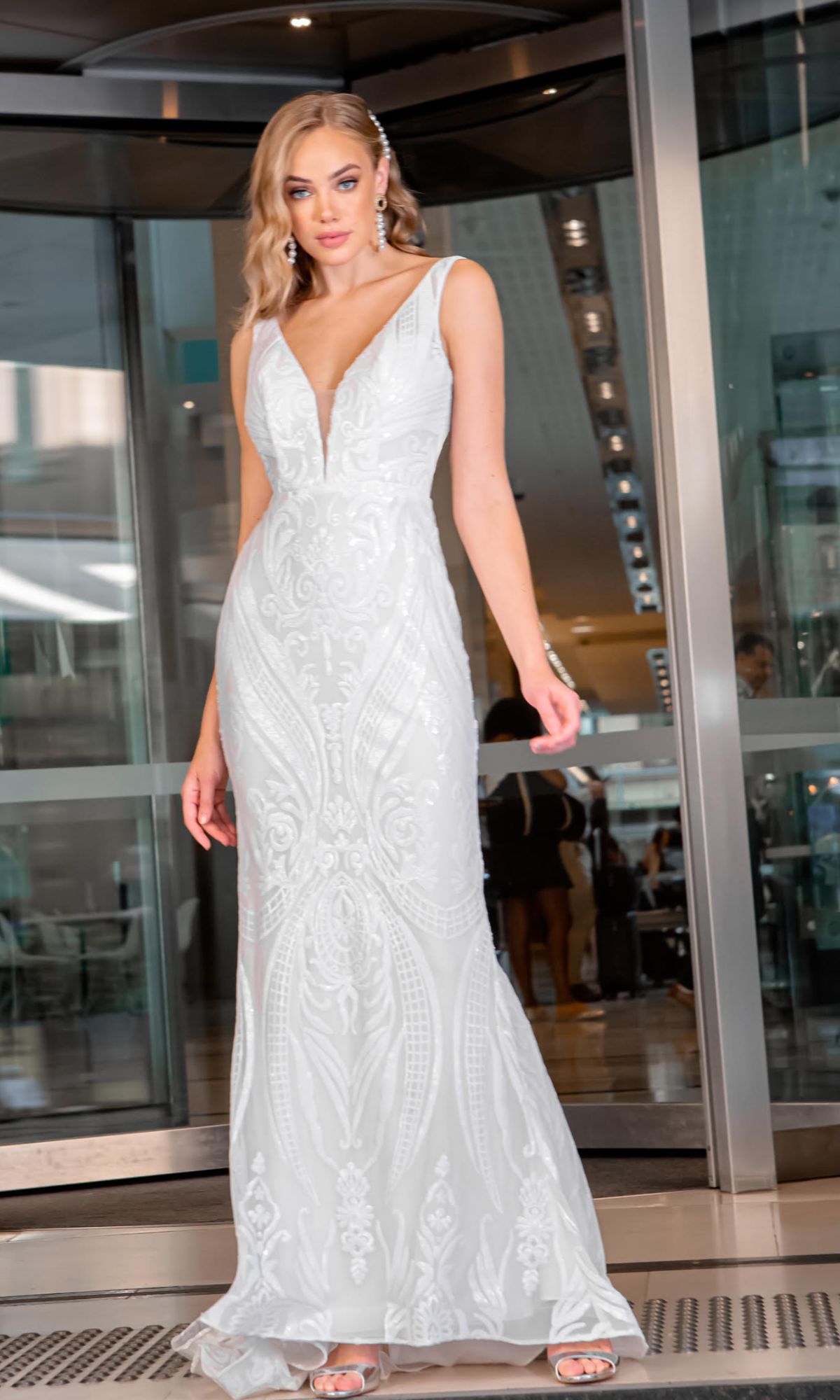 A woman wearing the White Lace Mermaid Wedding Gown: Tina Holly BA109 in white, featuring a deep V neckline, stands before glass doors.